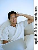 Man lying on sofa with laptop, hand on head, looking at camera