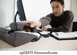 Male executive sitting at office desk, reaching for telephone Male executive sitting at office desk, reaching for telephone
