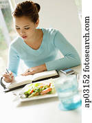 Young woman sitting at table, food next to her, writing in notebook Young woman sitting at table, food next to her, writing in notebook