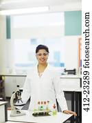 Portrait of smiling female teacher wearing protective eyewear, standing behind desk with microscope and test tubes in rack