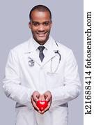 Your heart in right hands. Confident African doctor holding heart shape toy and smiling while standing against grey background