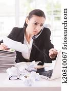 Busy office life. Frustrated young woman in formalwear talking on the telephone and holding documents while sitting at her working place
