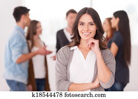 She is a team leader. Confident young woman holding hand on chin and smiling while group of people communicating on background She is a team leader. Confident young woman holding hand on chin and smiling while group of people communicating on background