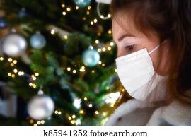Portrait of a young woman wearing protective face mask and looking sad for Covid-19 with Christmas tree on the background, coronavirus and Christmas concept