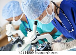 A female medical or scientific researcher or doctor using her microscope in a laboratory with her Asian colleague out of focus behind her.