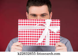 Present gift in hands of smiling man covering half face. closeup on happy man standing on red background