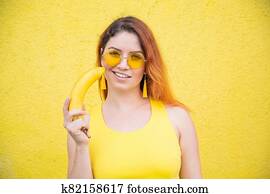 Portrait of a beautiful red-haired woman in a yellow dress and sunglasses holds a banana near sensual lips. Girl posing with fruit on a yellow background. Portrait of a beautiful red-haired woman in a yellow dress and sunglasses holds a banana near sensual lips. Girl posing with fruit on a yellow background.