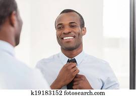 Confident about his look. Young African man adjusting his necktie while standing against mirror