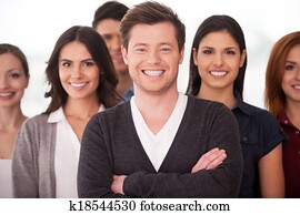He is a real team leader. Confident young man keeping arms crossed and smiling while group of people standing on background