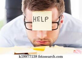 He needs help. Frustrated young man in formalwear with adhesive note on his forehead leaning his head at the table