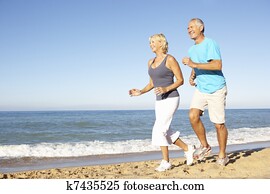 Senior Couple In Fitness Clothing Running Along Beach Senior Couple In Fitness Clothing Running Along Beach