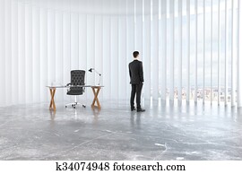 Businessman looks out the window in empty loft room with glassy table, leather chair and concrete floor