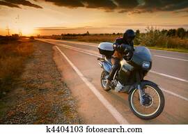 Biker and motorcycle on road at sunset Biker and motorcycle on road at sunset