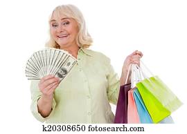 Senior shopaholic. Happy senior woman stretching out hand with paper currency and holing shopping bags while standing isolated on white background