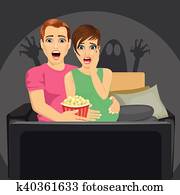 young couple watching horror movie at home sitting with popcorn on sofa