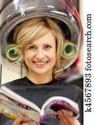 Delighted woman reading a magazine with hair curlers under a hairdryer in a hairdressing salon