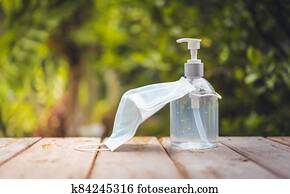 Face mask and white hand sanitizer or alcohol gel pump bottle on wooden top with green nature backgrounds Face mask and white hand sanitizer or alcohol gel pump bottle on wooden top with green nature backgrounds