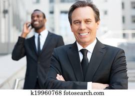 experte, in, geschaefts, world., sicher, älterer mann, in, formalwear, beibehaltung, verschränkte arme, und, lächeln, während, afrikanischer mann, sprechen, auf, dass, handy, in, dass, hintergrund