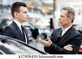 Discussing car features. Two people in formalwear talking to each other and gesturing while standing at the dealership