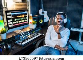 Young serious African musician in casualwear and headphones sitting in armchair