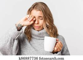 Close-up of beautiful sleepy woman with tatoos, drinking coffee from mug and rubbing eyes, standing over white background Close-up of beautiful sleepy woman with tatoos, drinking coffee from mug and rubbing eyes, standing over white background