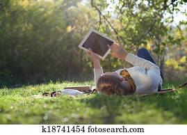 woman lying on bedding on green grass with ipad during picknic in the park woman lying on bedding on green grass with ipad during picknic in the park