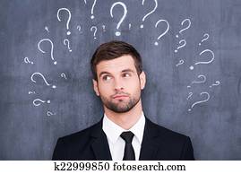 So many questions! Confused young man standing against blackboard with question marks