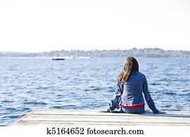 Girl sitting alone on dock by lake, looking out over water.