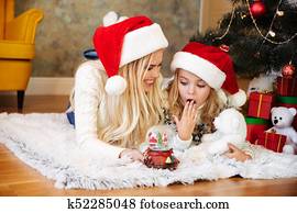 Surprised little girl getting snow ball toy from her mother while lying on the floor near Christmas tree