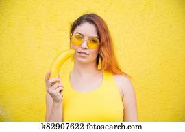 Portrait of a beautiful red-haired woman in a yellow dress and sunglasses holds a banana near sensual lips. Girl posing with fruit on a yellow background. Portrait of a beautiful red-haired woman in a yellow dress and sunglasses holds a banana near sensual lips. Girl posing with fruit on a yellow background.