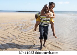 Young Man Giving Woman Piggyback Along Shoreline Of Beach Young Man Giving Woman Piggyback Along Shoreline Of Beach