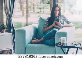 Lovely message from him. Beautiful young smiling woman looking at her smart phone while sitting in a big comfortable chair at home