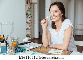 Smiling woman painter holding paintbrush and thinking at the table Smiling woman painter holding paintbrush and thinking at the table