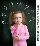 Child in eyeglasses standing near school blackboard with many question marks