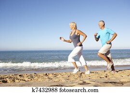Senior Couple In Fitness Clothing Running Along Beach Senior Couple In Fitness Clothing Running Along Beach