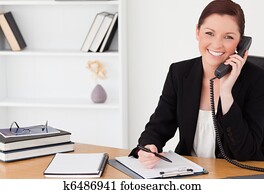 Pretty red-haired woman in suit writing on a notepad and phoning while sitting in an office