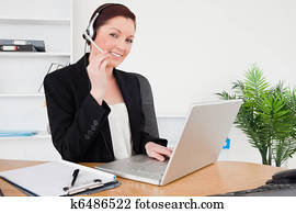Young attractive red-haired female in suit typing on her laptop and using headphones while sitting in an office