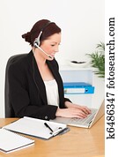 Young attractive red-haired woman in suit typing on her laptop and using headphones while sitting in an office