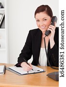 Young good looking red-haired woman in suit writing on a notepad and phoning while sitting in an office