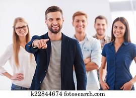 Join our team! Beautiful young woman showing her thumb up and smiling while group of happy young people standing on background and smiling