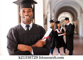 Confident graduate. Happy African man in graduation gowns holding diploma and smiling while his friends standing in the background Confident graduate. Happy African man in graduation gowns holding diploma and smiling while his friends standing in the background