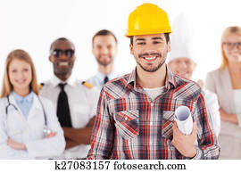 When I grow up I will be an engineer. Confident young man in hardhat holding blueprint and smiling while group of people in different professions standing in the background