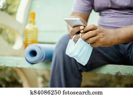 Man using mobile with mask in hand at park during morning yoga or workout with mat - concept of coronavirus or covid-19 advice, protection or to prevent from spreading by wearing mask at outdoor. Man using mobile with mask in hand at park during morning yoga or workout with mat - concept of coronavirus or covid-19 advice, protection or to prevent from spreading by wearing mask at outdoor.