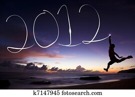 happy new year 2012. young man jumping and drawing 2012 by flashlight in the air on the beach before sunrise