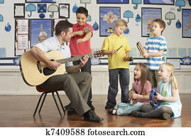 Male Teacher Playing Guitar With Pupils Having Music Lesson In Classroom Male Teacher Playing Guitar With Pupils Having Music Lesson In Classroom