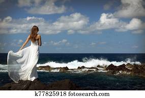 Young bride in light white wedding dress standing on sea rock shore and looking at ocean. Back view Young bride in light white wedding dress standing on sea rock shore and looking at ocean. Back view