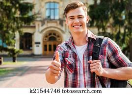 Thumb up for success! Handsome male student showing his thumb up and smiling while standing outdoors with university building in the background