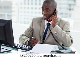 Smiling entrepreneur making a phone call while looking at his computer in his office Smiling entrepreneur making a phone call while looking at his computer in his office