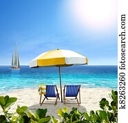 Beautiful beach with white sand, two chairs, white-yellow umbrella and a sailing boat in the sea.