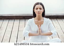 In balance with herself. Beautiful young woman in white clothing sitting in lotus position and keeping hands clasped while meditating outdoors